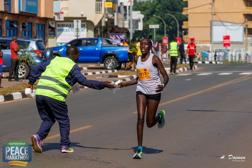 A race official in a yellow safety vest hands a water bottle to a female runner during a street marathon event in an urban area