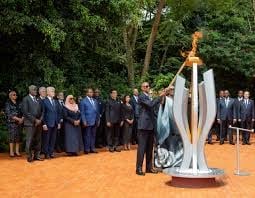 Group of formally dressed people gathered around a large modern white sculpture with a flame on top in an outdoor setting