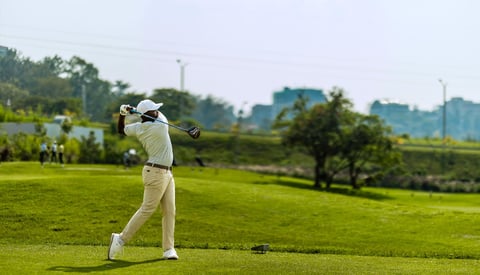 Golfer in beige attire swinging on a lush green golf course with trees and distant landscape in background