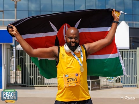 Athlete in yellow race bib holding Kenyan flag overhead after finishing marathon, wearing gold medals
