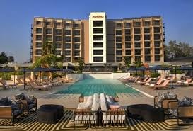 Modern resort hotel with beige facade overlooking a large swimming pool surrounded by lounge chairs and umbrellas