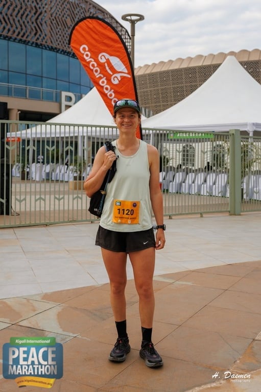 Female runner wearing race bib 1120 at Peace Marathon event, posing in front of large orange parasol and white tent structure with modern building in background