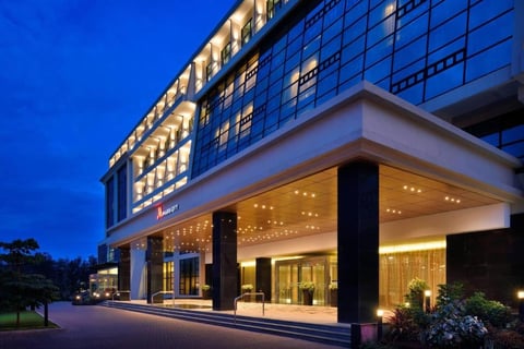 Modern hotel building illuminated at dusk with glass facade, columned entrance, and landscaping against blue twilight sky