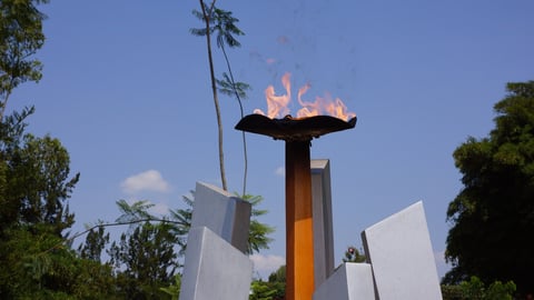 Eternal flame monument with burning torch atop a tall pillar surrounded by white panels and green trees against a blue sky